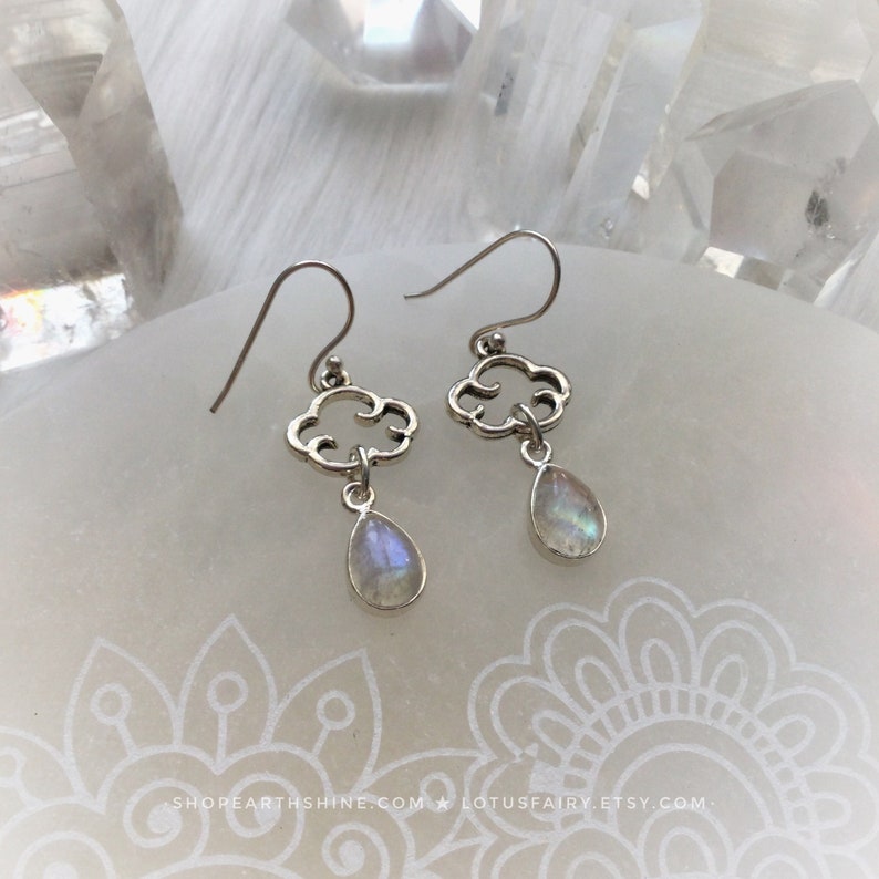 May include: Silver cloud-shaped earrings with teardrop-shaped, iridescent moonstone pendants. The earrings have a hook closure and are displayed on a white surface with a floral design. The earrings are a delicate and elegant accessory.