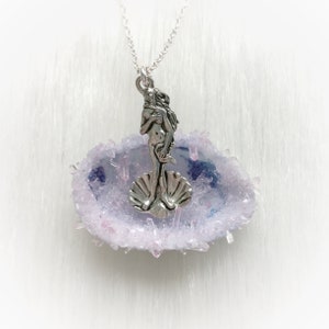 May include: A silver pendant necklace featuring a depiction of the Venus de Milo standing on a seashell, set within a purple and pink agate slice.