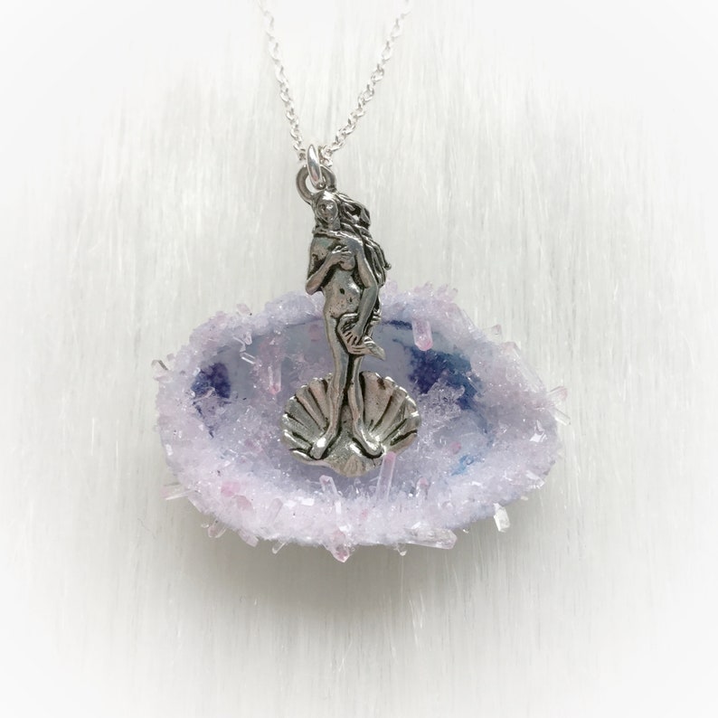 May include: A silver pendant necklace featuring a depiction of the Venus de Milo standing on a seashell, set within a purple and pink agate slice.