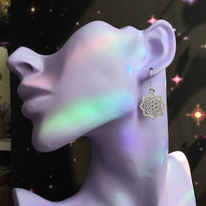 May include: A silver earring with a geometric flower of life design is displayed on a lavender bust. The earring hangs from a simple hook. The background is dark with a pattern of stars and colorful light reflections.