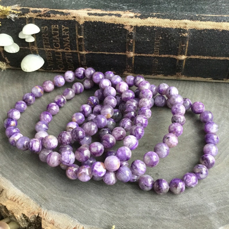 May include: Several purple charoite gemstone bracelets. The bracelets are made of round, polished beads with a marbled appearance. The background includes a vintage book and decorative mushrooms, creating a natural, earthy aesthetic.