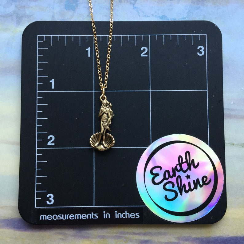 May include: Gold necklace with a bronze pendant depicting the Birth of Venus sculpture. The pendant is about 1 inch tall and the necklace is about 18 inches long. The necklace is on a black background with a ruler and a sticker that says "Earth Shine".