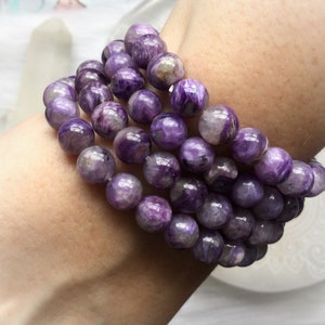 May include: A stack of purple beaded bracelets. The beads are round and have a marbled appearance with shades of lavender, violet, and white. The bracelets are worn on a wrist, showcasing the jewelry's color and texture.