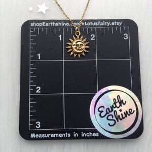 May include: Gold sun and moon necklace with a face on the moon. The necklace is 1 inch in diameter and is shown on a black grid with measurements in inches.