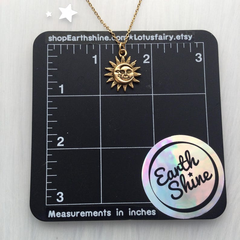 May include: Gold sun and moon necklace with a face on the moon. The necklace is 1 inch in diameter and is shown on a black grid with measurements in inches.