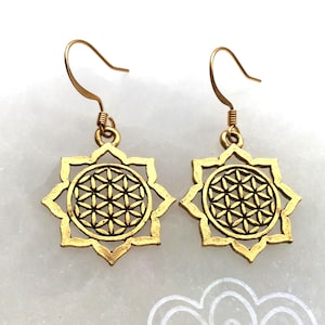 May include: A pair of gold-coloured earrings. Each earring features a flower-like design with a geometric pattern in the centre. The earrings have a hook closure and are displayed on a white surface.