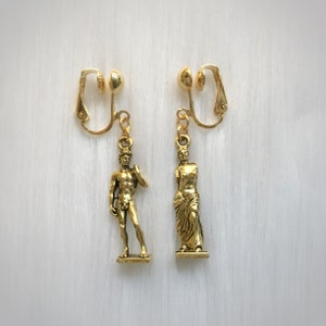 May include: A pair of gold-colored clip-on earrings. Each earring features a small, detailed sculpture. One depicts a male figure, the other a female figure. The earrings are displayed on a white, textured surface.