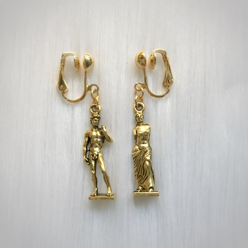 May include: A pair of gold-colored clip-on earrings. Each earring features a small, detailed sculpture. One depicts a male figure, the other a female figure. The earrings are displayed on a white, textured surface.