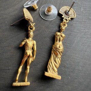 May include: Gold-toned dangle earrings featuring classical figures. One earring depicts a male figure, the other a draped female form. The earrings have a post-back closure with clear plastic earring backs. The earrings are set against a dark background.