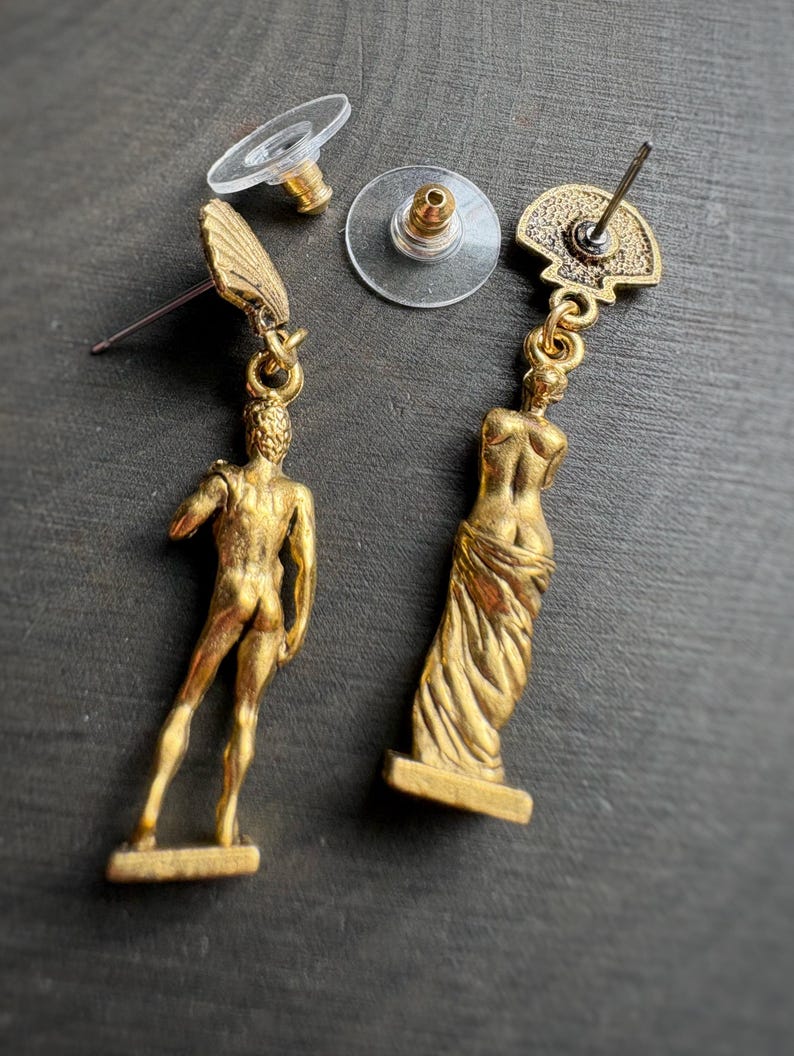 May include: Gold-toned dangle earrings featuring classical figures. One earring depicts a male figure, the other a draped female form. The earrings have a post-back closure with clear plastic earring backs. The earrings are set against a dark background.