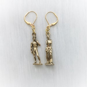 May include: A pair of gold-colored earrings. One earring features a miniature statue of David, and the other depicts a classical statue. Both earrings have lever-back closures and are suspended from gold-toned ear wires.
