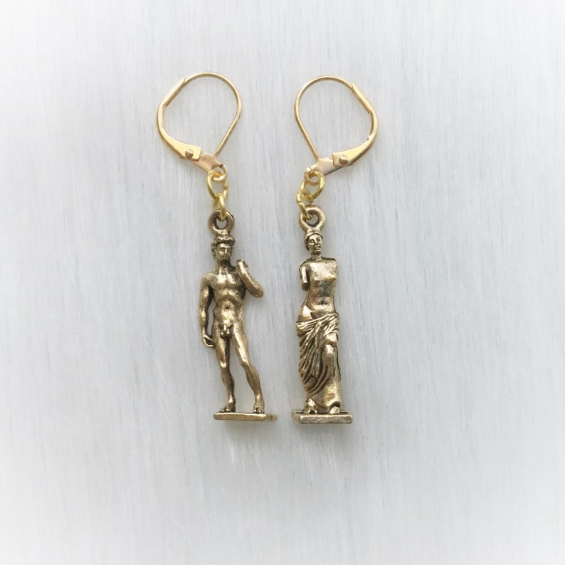 May include: A pair of gold-colored earrings. One earring features a miniature statue of David, and the other depicts a classical statue. Both earrings have lever-back closures and are suspended from gold-toned ear wires.