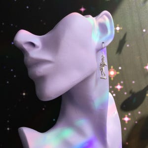 May include: A silver-colored earring featuring a small, detailed figure of a man. The earring hangs from a simple hook. The earring is displayed on a lavender-colored bust. The background is dark with star-like shapes.