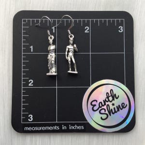 May include: Silver-toned dangle earrings featuring miniature sculptures of classical figures. One earring depicts a draped statue, the other a nude male figure. The earrings are displayed on a black surface with inch measurements.