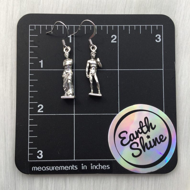 May include: Silver-toned dangle earrings featuring miniature sculptures of classical figures. One earring depicts a draped statue, the other a nude male figure. The earrings are displayed on a black surface with inch measurements.