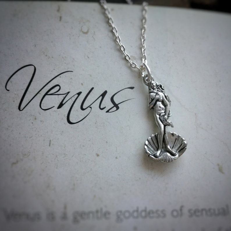 May include: Silver necklace with a pendant featuring a depiction of the Venus de Milo statue. The pendant is shaped like a seashell and the statue is standing on top of it. The word "Venus" is written in cursive on the background.