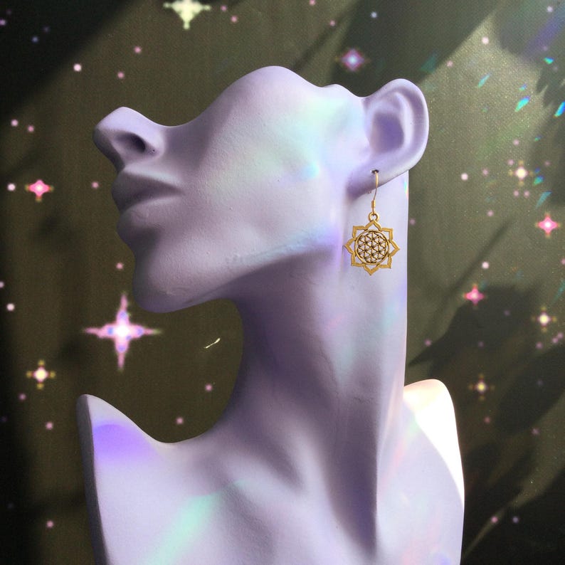 May include: Gold-colored earrings with a geometric starburst design are displayed on a lavender bust. The earrings have a delicate, openwork pattern. The background is a dark, shimmering color with star-shaped highlights.