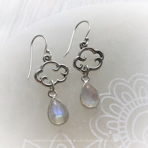 May include: A pair of silver earrings featuring a cloud design with a teardrop-shaped, iridescent gemstone pendant. The earrings have a hook closure and are displayed on a white surface. The earrings are from the Lotus Fairy Etsy shop.