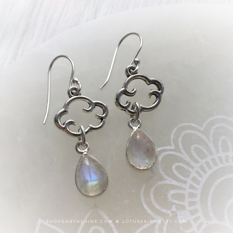 May include: A pair of silver earrings featuring a cloud design with a teardrop-shaped, iridescent gemstone pendant. The earrings have a hook closure and are displayed on a white surface. The earrings are from the Lotus Fairy Etsy shop.