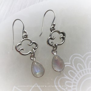 May include: A pair of silver earrings featuring a cloud-shaped design with a teardrop-shaped, iridescent gemstone pendant. The earrings have a hook closure and are displayed on a white surface.