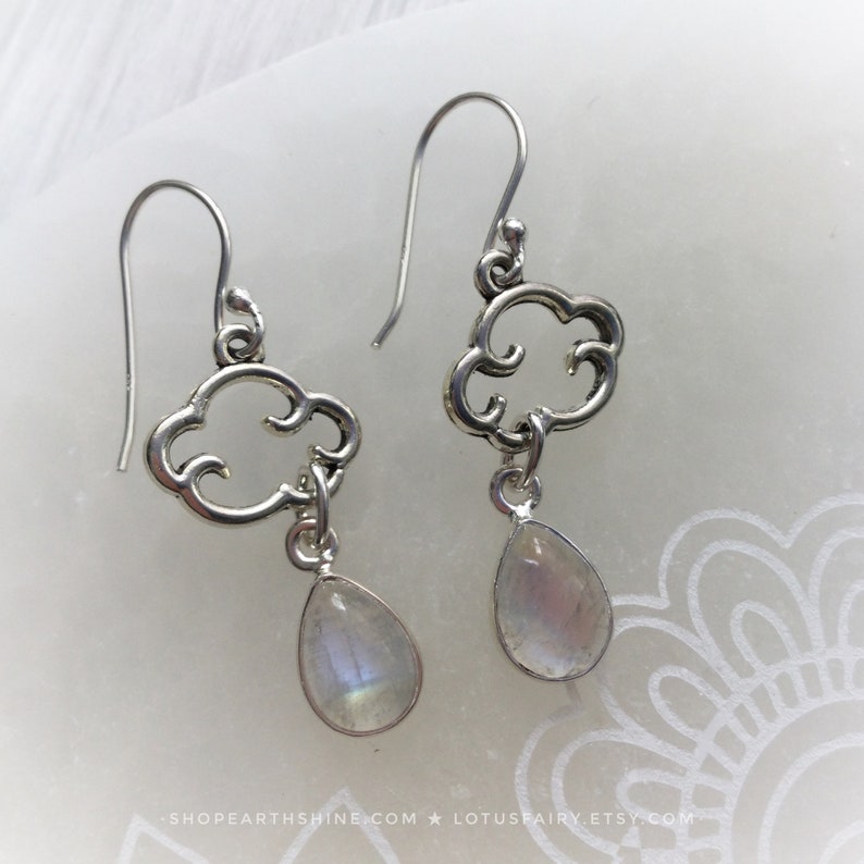 May include: A pair of silver earrings featuring a cloud-shaped design with a teardrop-shaped, iridescent gemstone pendant. The earrings have a hook closure and are displayed on a white surface.