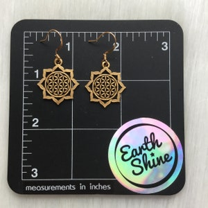 May include: A pair of gold-colored earrings with a flower of life design. The earrings are displayed on a black surface with measurements in inches. The earrings are approximately 1 inch in diameter. The Earth Shine logo is visible.