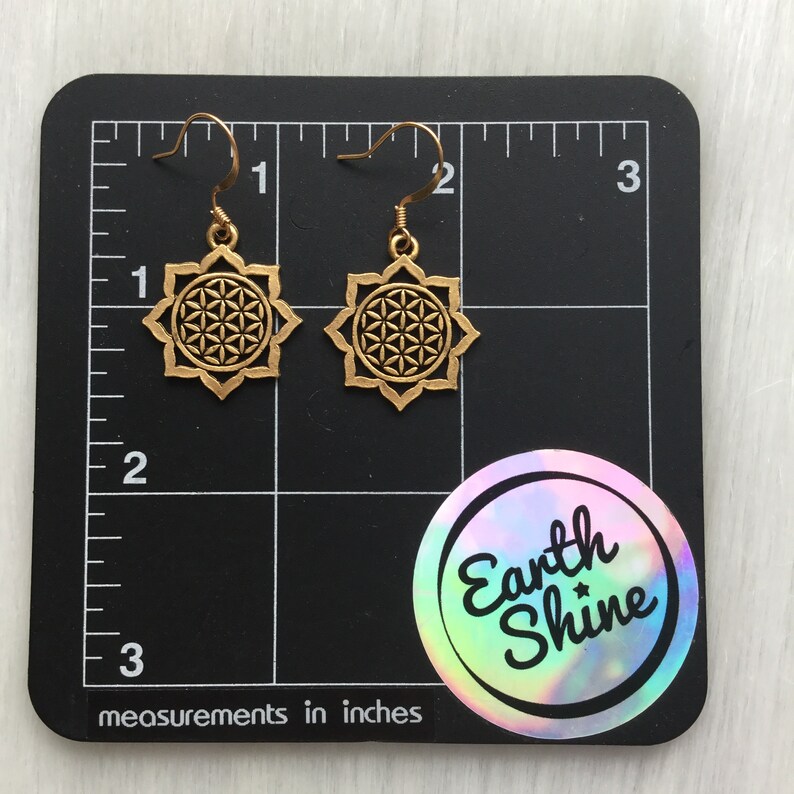 May include: A pair of gold-colored earrings with a flower of life design. The earrings are displayed on a black surface with measurements in inches. The earrings are approximately 1 inch in diameter. The Earth Shine logo is visible.