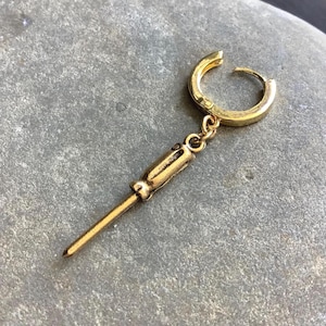 May include: A gold-coloured earring with a screwdriver charm. The earring has a small hoop and a detailed screwdriver pendant. The metal has a slightly aged appearance. The earring is likely made of brass or a similar alloy.