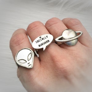 Könnte beinhalten: Drei silberne Ringe mit unterschiedlichen Designs. Der erste Ring zeigt einen silbernen Alienkopf. Der zweite Ring ist eine silberne Sprechblase mit dem Text "Socially Awkward". Der dritte Ring ist ein silberner Planet mit einem Ring darum.