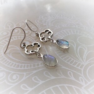 May include: Silver-toned dangle earrings. Each earring features a decorative cloud-shaped top with a teardrop-shaped, iridescent gemstone. The gemstones display a range of colors, including blue, purple, and white. The earrings are designed with a hook closure.