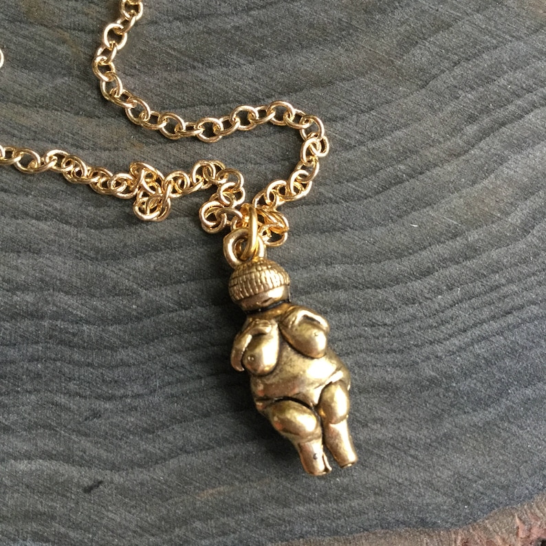 May include: A gold necklace with a pendant shaped like a stylized female figure. The figure is depicted with large breasts and a rounded belly.