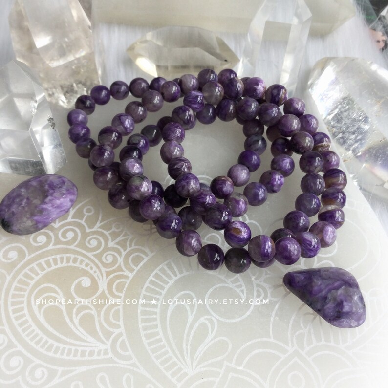 May include: Several strands of purple beaded bracelets and two polished stones. The beads and stones have a marbled appearance with shades of purple and white. The bracelets are arranged in a circular pattern. Clear quartz crystals are in the background.