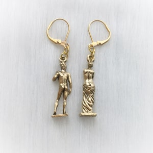 May include: A pair of gold-colored dangle earrings. One earring features a miniature statue of David, and the other a statue of a draped figure. Both statues are suspended from gold-tone lever-back closures.