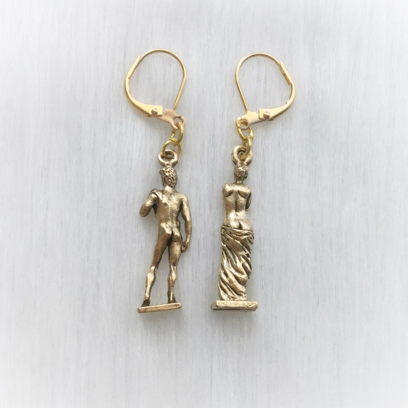 May include: A pair of gold-colored dangle earrings. One earring features a miniature statue of David, and the other a statue of a draped figure. Both statues are suspended from gold-tone lever-back closures.
