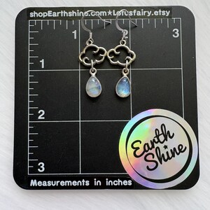 May include: Silver cloud-shaped earrings with teardrop-shaped, iridescent gemstones. The earrings are displayed on a black grid with inch measurements. The text "shopEarthshine.com" and "Earth Shine" are visible.