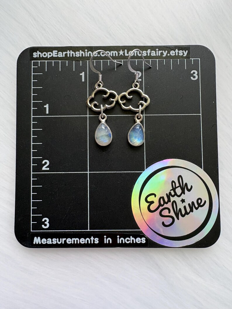 May include: Silver cloud-shaped earrings with teardrop-shaped, iridescent gemstones. The earrings are displayed on a black grid with inch measurements. The text "shopEarthshine.com" and "Earth Shine" are visible.