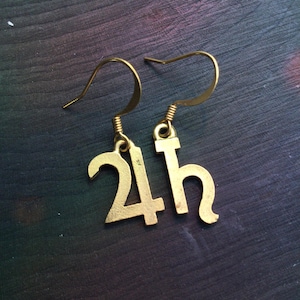 May include: A pair of gold-colored earrings. Each earring features a hook and a charm in the shape of the numbers "24h". The earrings are suspended from a coiled wire. The background is a dark, textured surface.