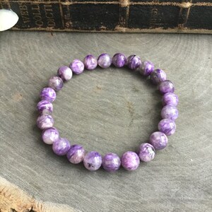 May include: A beaded bracelet featuring round, polished beads in shades of purple and white. The bracelet is made of natural stone and is displayed on a gray surface. The beads have a marbled appearance.