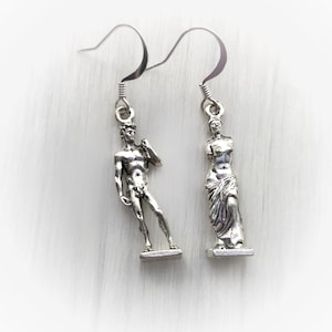 May include: Silver-toned dangle earrings featuring miniature sculptures of classical figures. One earring depicts a standing male figure, the other a draped female form. The earrings have a simple hook design and are set against a white background.