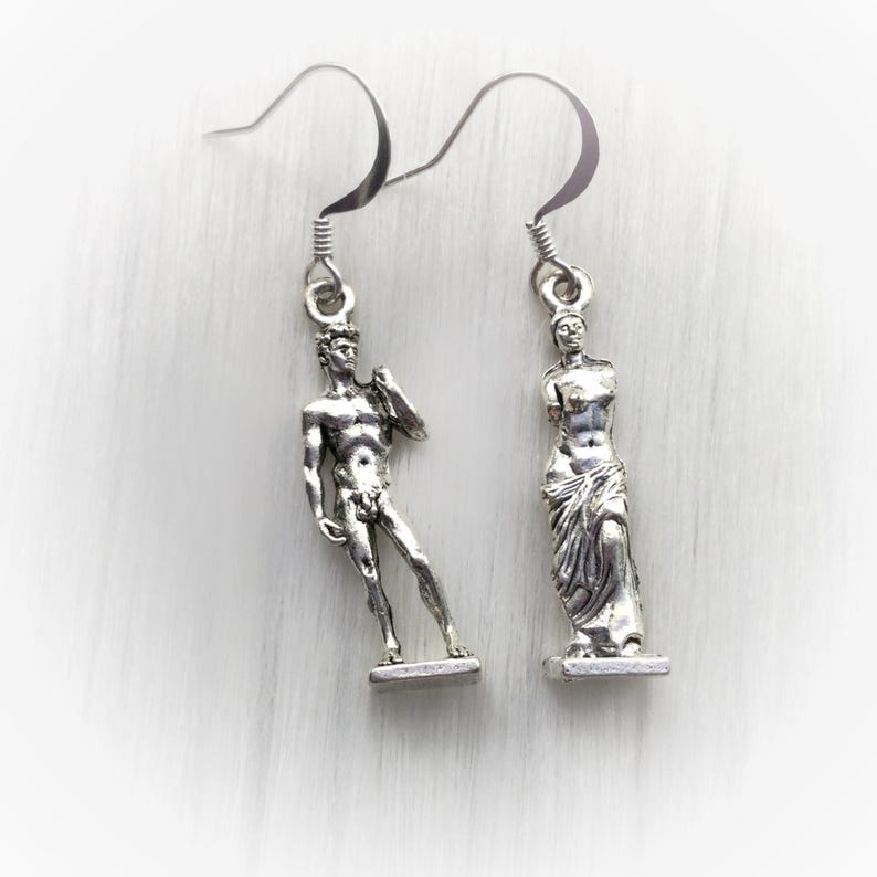 May include: Silver-toned dangle earrings featuring miniature sculptures of classical figures. One earring depicts a standing male figure, the other a draped female form. The earrings have a simple hook design and are set against a white background.
