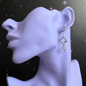 May include: Silver cloud earrings with a teardrop-shaped, clear gemstone. The earrings are displayed on a lavender-colored bust against a black background with white stars.
