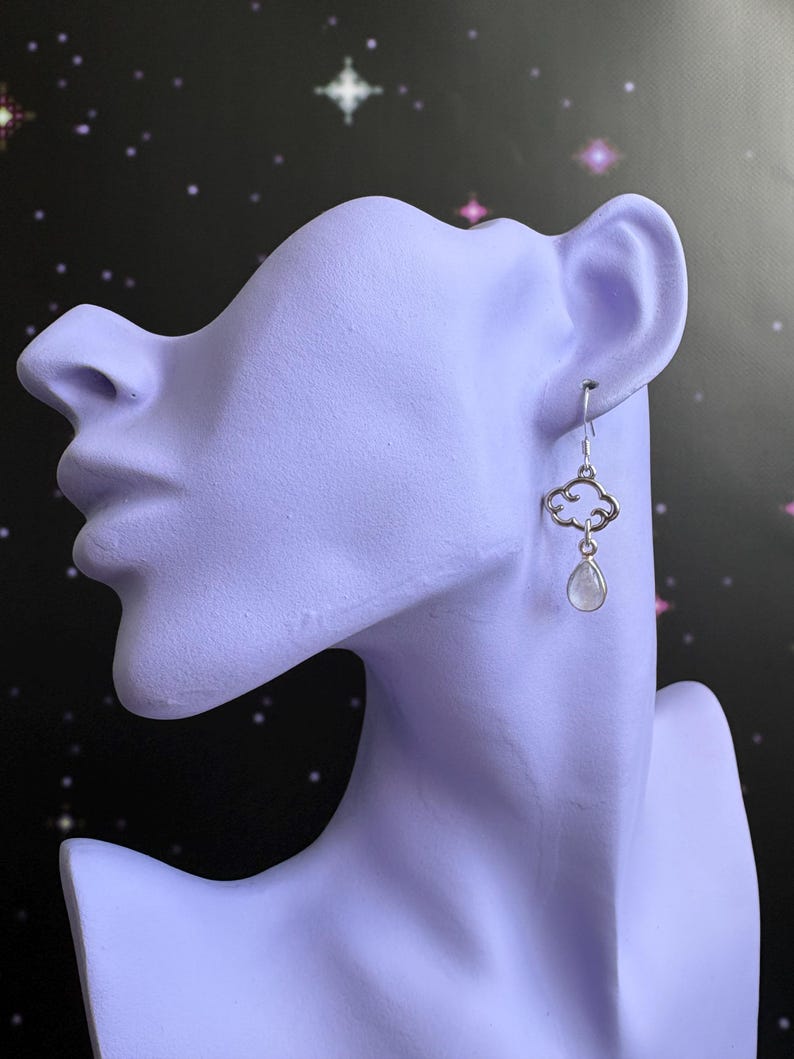 May include: Silver cloud earrings with a teardrop-shaped, clear gemstone. The earrings are displayed on a lavender-colored bust against a black background with white stars.