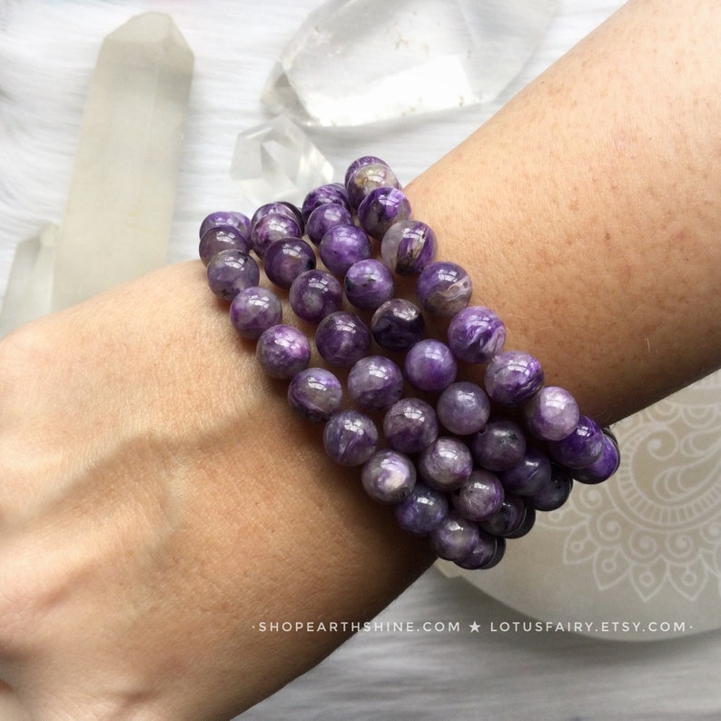 May include: A close-up of several bracelets made of round, purple beads. The beads have a marbled appearance with lighter purple and white swirls. The bracelets are stacked on a person's wrist. The background includes crystals and the text "SHOPEARTHSHINE.COM * LOTUSFAIRY.ETSY.COM".