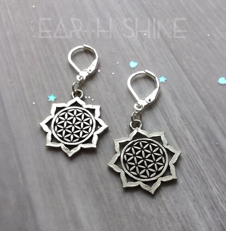 May include: A pair of silver-colored earrings. Each earring features a flower-like design with a central geometric pattern. The earrings have lever-back closures and are displayed on a light gray surface.