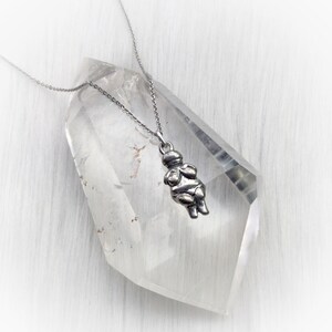 May include: A silver necklace with a pendant shaped like a stylized female figure. The pendant is hanging from a silver chain and is resting on a clear quartz crystal.