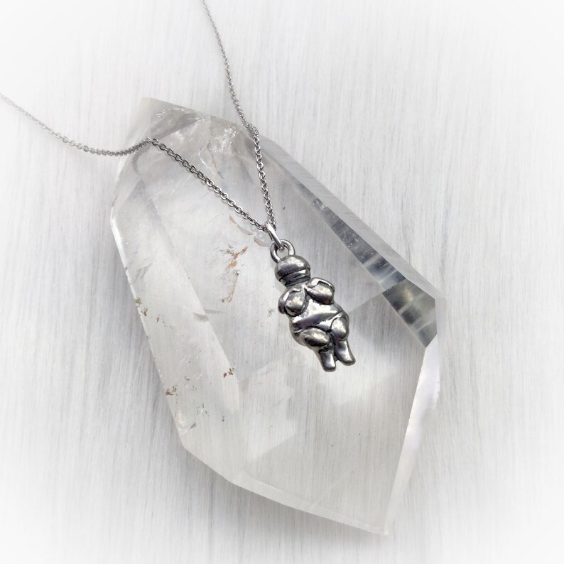 May include: A silver necklace with a pendant shaped like a stylized female figure. The pendant is hanging from a silver chain and is resting on a clear quartz crystal.