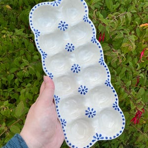 May include: A white ceramic egg tray with twelve circular indentations, featuring a hand-painted blue floral pattern along the scalloped edges and a series of small blue flower motifs running down the center of the rectangular serving piece.