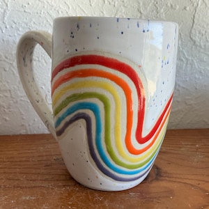 Pode incluir: Uma caneca de cerâmica branca com acabamento brilhante e textura salpicada. O exterior é decorado com um padrão de arco-íris ondulado em relevo nas cores vermelho, laranja, amarelo, verde, azul e roxo. A caneca inclui uma alça curva.