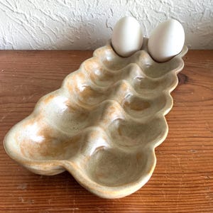 May include: A ceramic egg holder with a wavy design, holding two white eggs. The holder is a light green color with brown accents, and has space for several eggs. The handmade egg tray sits on a wooden surface.