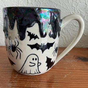 Puede incluir: Una taza de cerámica con base color crema que presenta ilustraciones negras pintadas a mano de un fantasma, murciélagos y arañas. El borde superior tiene un esmalte negro iridiscente grueso que gotea por los lados de la pieza.